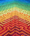 Closeup image, taken at an angle, of a crocheted shawl with geometric patterns, red and irange near the camera, going through yellow and green to blue.